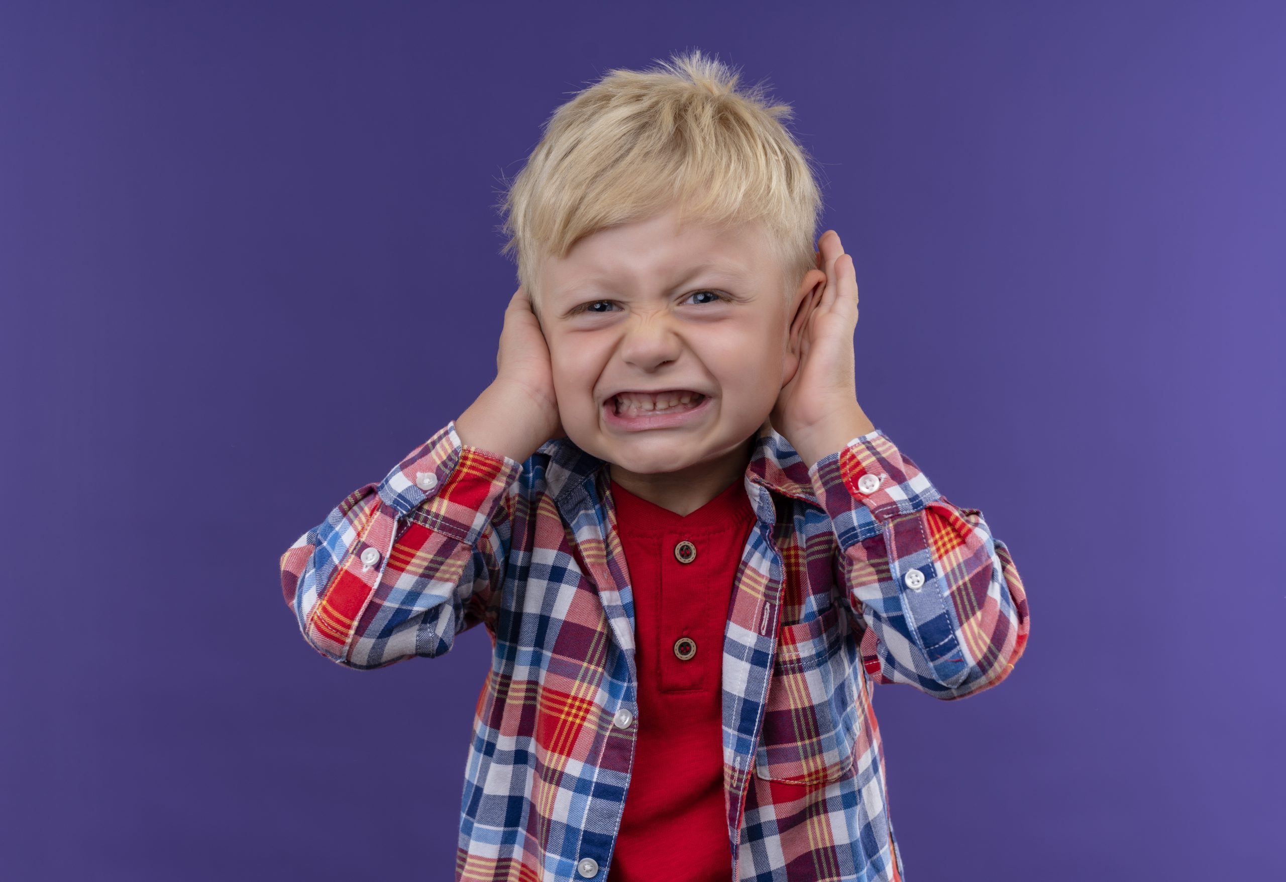 an angry little boy with blonde hair wearing checked shirt keeping hands on ears on a purple background
